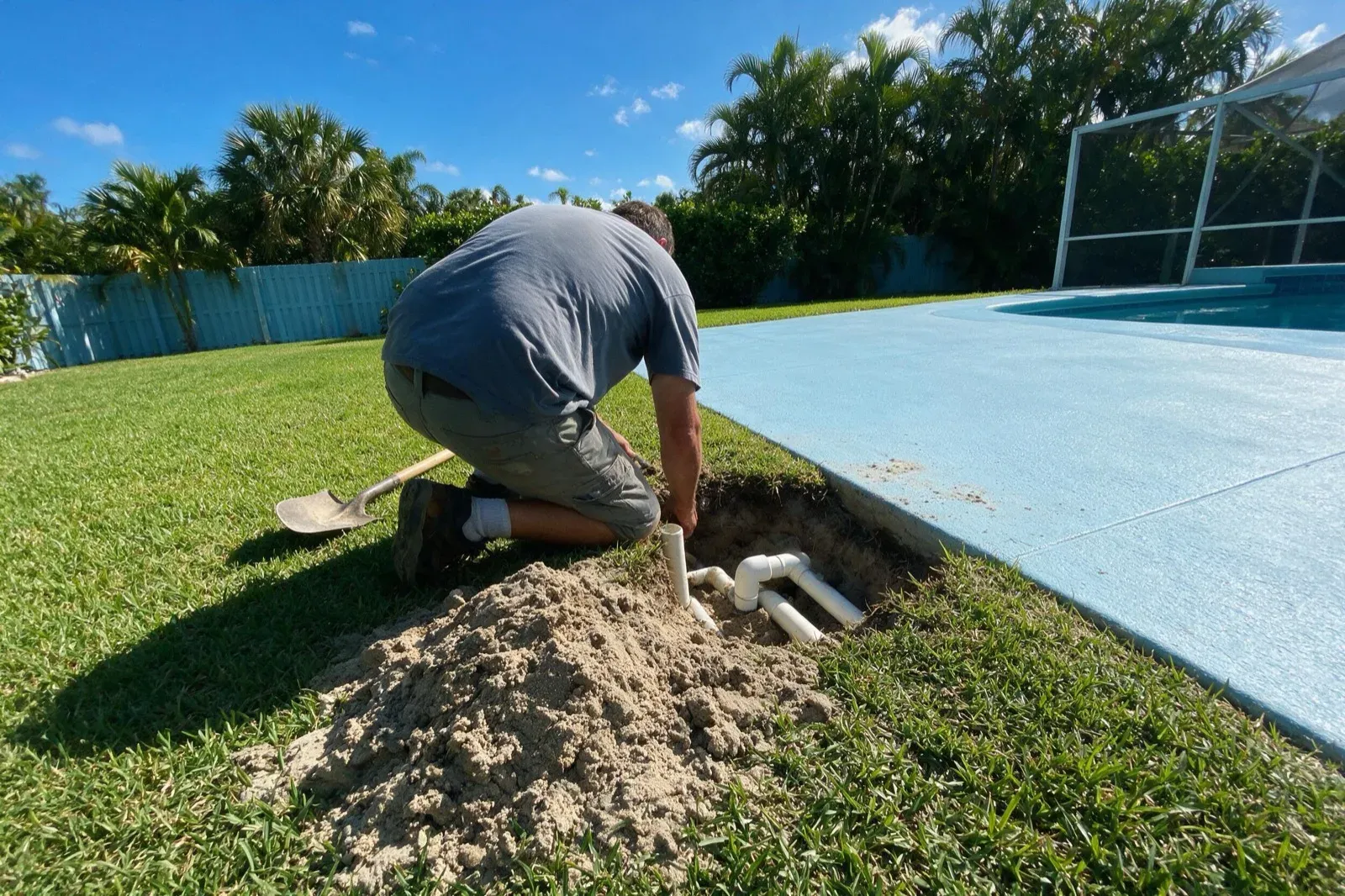 Technician inspecting exposed pool plumbing in an access dig beside the deck