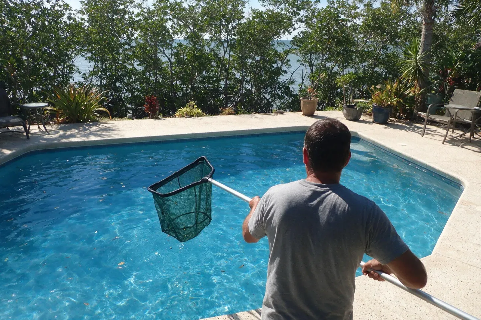 Technician skimming a Marco Island pool with coastal water beyond the deck